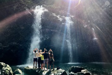 Guests enjoying a waterfall view from rocks in Maui. The sun is beaming down on them.